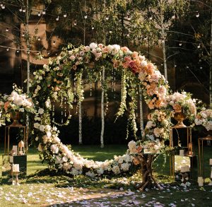 Outdoor garden wedding ceremony arch designed by Tiffany Siegel, adorned with a lush array of roses and hanging blooms, complemented by delicate string lights, and framed by nature's greenery, setting the stage for an enchanting matrimonial setting.