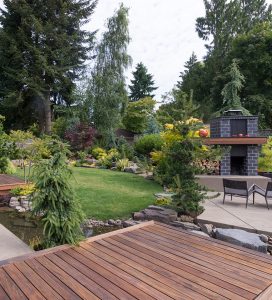 Expertly crafted garden design by Tiffany Siegel, showcasing a variety of textured foliage and vibrant plants, with a neatly edged lawn, complemented by a sleek wooden deck and a modern outdoor fireplace, creating a perfect blend of nature and contemporary outdoor living.