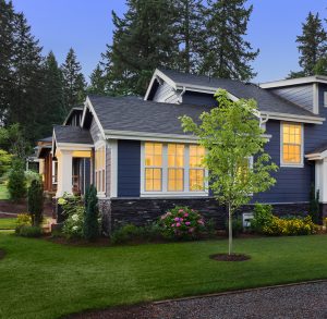Charming residential home exterior with a vibrant outdoor landscape designed by Tiffany Siegel, featuring a neatly manicured lawn, an array of lush shrubs and flowering plants, and a young, thriving tree, enhancing the home's welcoming appeal.