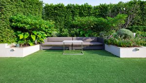 Minimalist and serene outdoor lounge area created by Tiffany Siegel, with a modern, plush outdoor sofa set against a backdrop of a meticulously trimmed privacy hedge and vibrant garden planters, offering a peaceful green retreat in an urban setting.