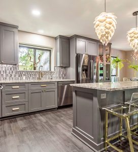 Chic kitchen design by Tiffany Siegel, featuring gray cabinetry with brass hardware, an intricate tiled backsplash, and elegant pendant lighting. The space is finished with a polished kitchen island, bar stools with gold accents, and a view of the outdoors.