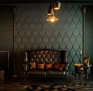 Vintage-inspired living room design by Tiffany Siegel, highlighted by a classic tufted leather sofa, rich damask-patterned wallpaper, ambient pendant lighting casting a warm glow, and antique wooden side tables, creating an atmosphere of timeless elegance.
