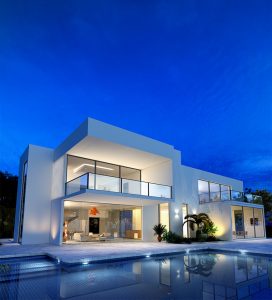 Modern white two-story home lit at night above a swimming pool